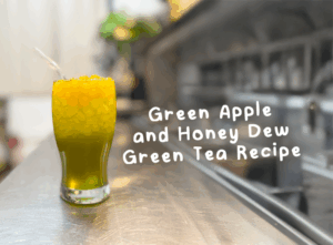 A refreshing Green Apple and Honey Dew green tea recipe served in a tall glass, topped with orange popping boba, placed on a stainless steel counter with a blurred cafe background.