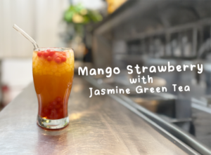A tall glass of Mango Strawberry with Jasmine Green Tea, topped with crushed ice and strawberry popping boba, placed on a metal surface.