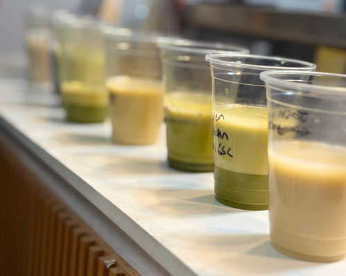 Row of clear plastic cups filled with various matcha and milk tea drink samples on a white counter