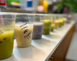 A row of labeled plastic cups with various milk tea samples lined up on a counter, including a jasmine crème blend in focus.