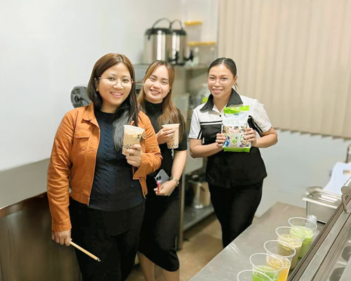 Three women smiling during a ProPossmei drink ideation session, holding milk tea and product samples inside a beverage test kitchen.