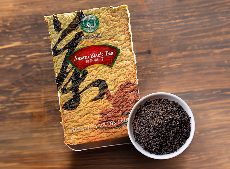 A package of POSSMEI Assam Black Tea placed beside a white bowl filled with loose Assam black tea leaves, on a wooden surface.