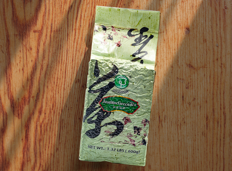A sealed pack of POSSMEI Jasmine Green Tea placed on a wooden surface with soft sunlight and shadows.
