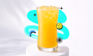A refreshing Kumquat Lemon Pop Recipe drink in a tall curvy glass with ice, placed on a white podium with a bright aesthetic background