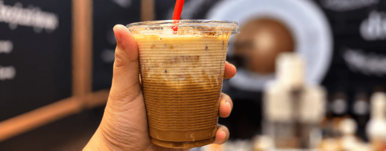 A hand holding a creamy iced coffee in a clear cup, showcasing its layered appearance and appealing presentation.