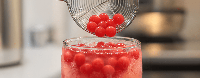 Red popping boba being added to a refreshing iced fruit drink, highlighting the drink’s visual appeal and customizable toppings.