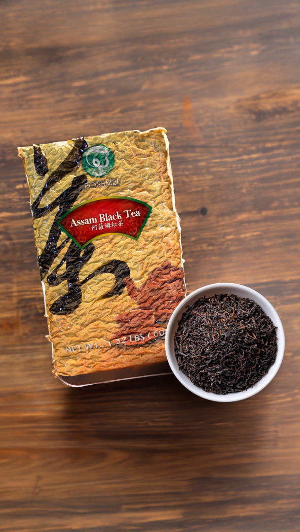 A package of POSSMEI Assam Black Tea placed beside a white bowl filled with loose Assam black tea leaves on a wooden surface.
